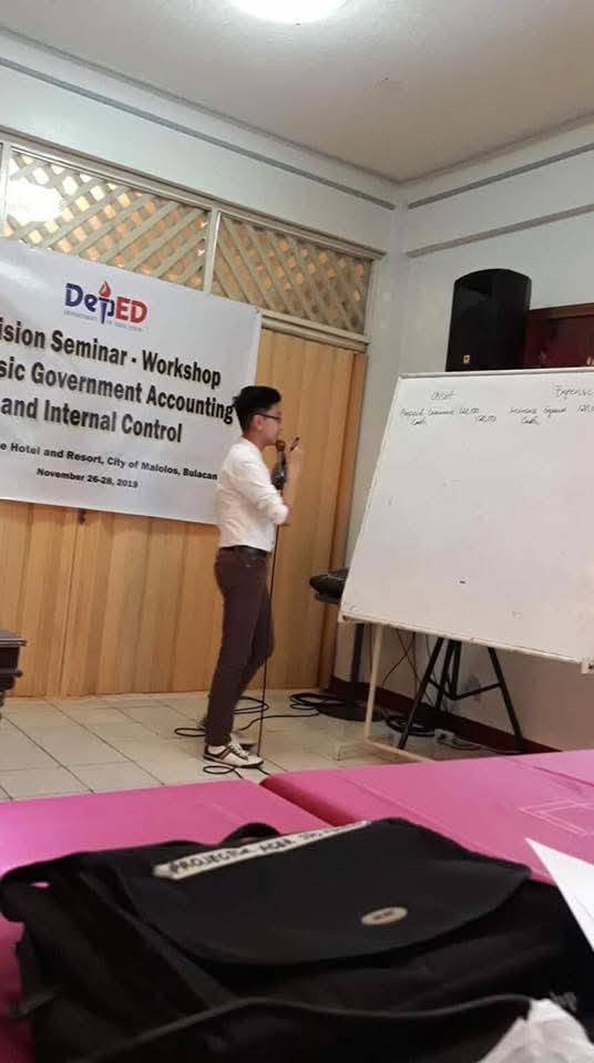 Jayson delivering the DepEd Division bookkeeping seminar beside the event banner in Malolos City