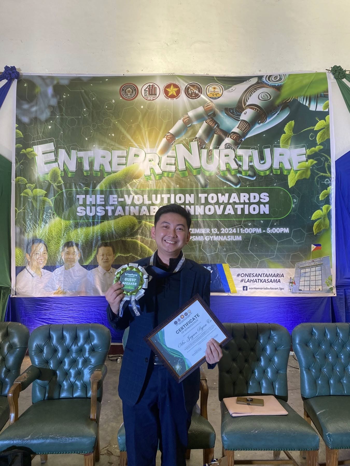 Jayson holding his guest speaker rosette and certificate of appreciation in front of the EntrepreNurture banner