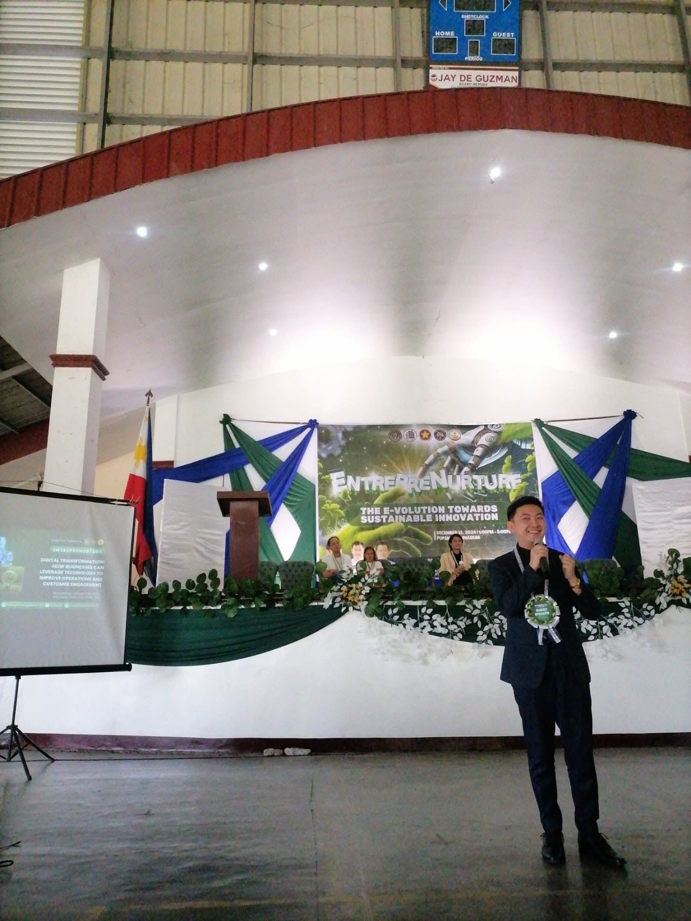 Jayson Ryan Co speaking with a microphone beside a Digital Transformation slide at the EntrepreNurture event in the PUP Santa Maria gymnasium