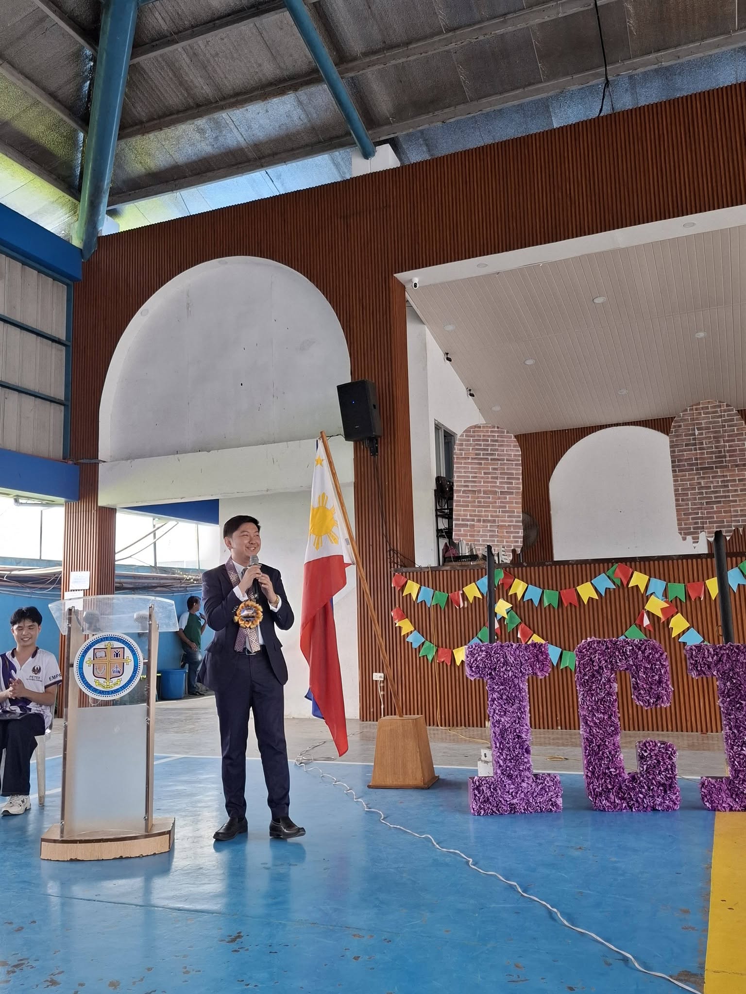 Jayson smiling as he addresses the audience beside the podium and the Philippine flag