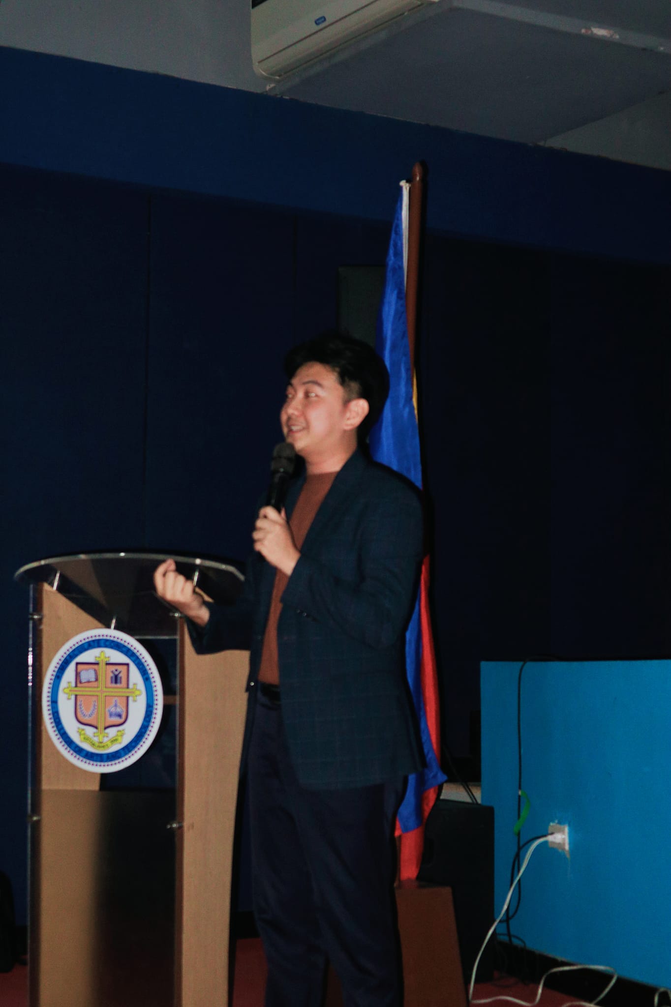 Jayson speaking at the ICI College podium with the Philippine flag behind him