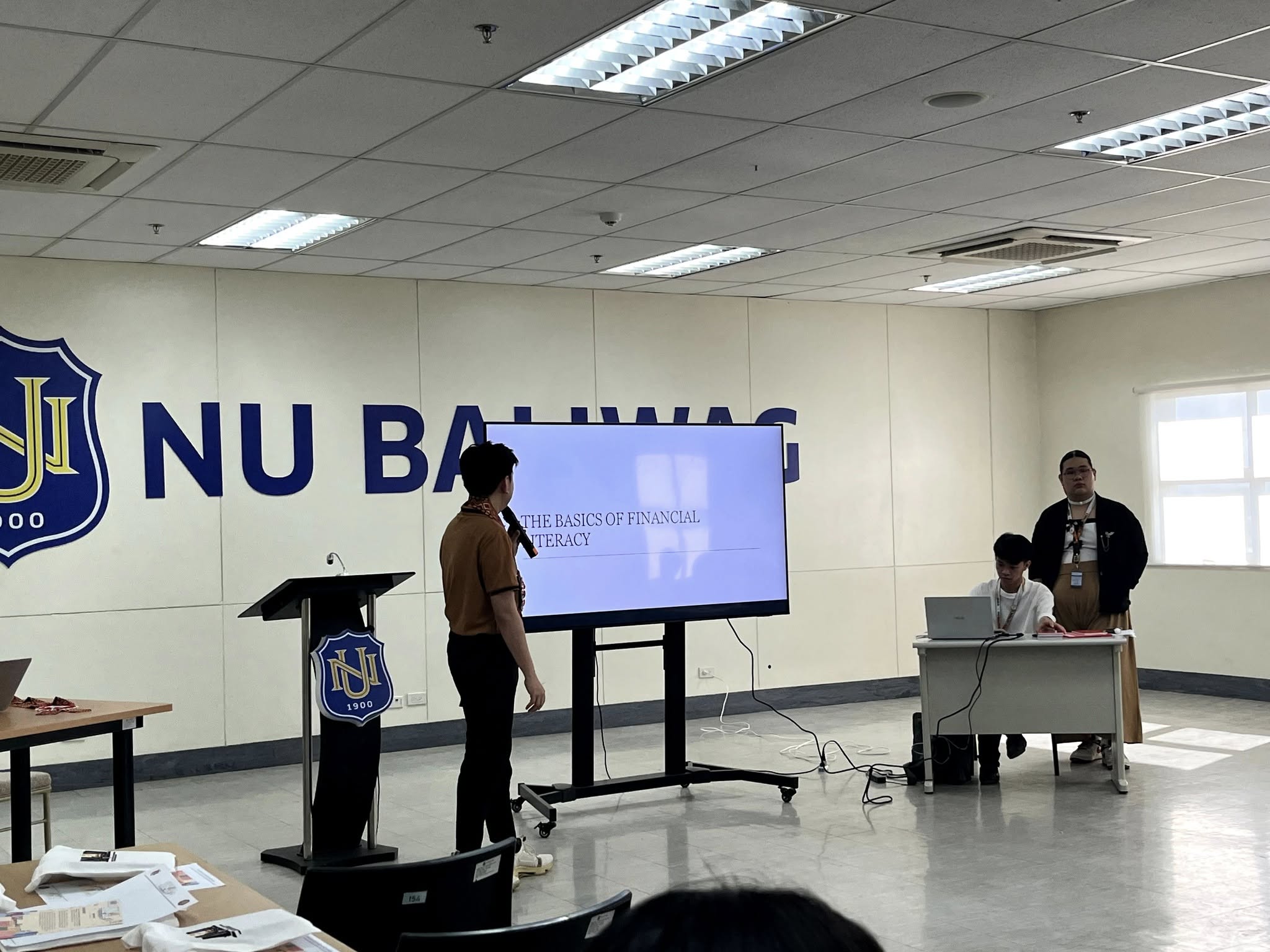 Jayson opening The Basics of Financial Literacy session beside the NU Baliwag wall logo