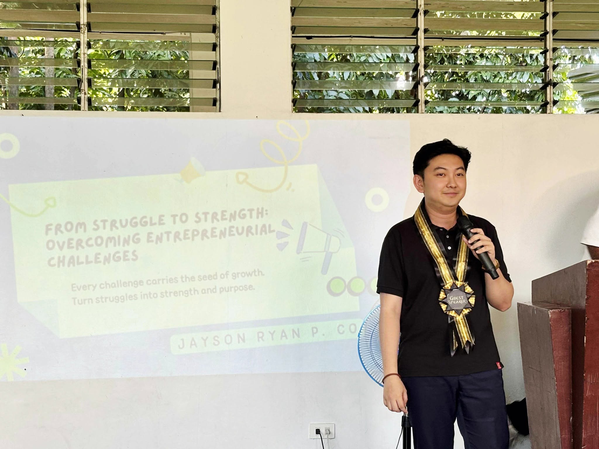 Jayson Ryan Co speaking with a microphone beside a slide titled From Struggle to Strength: Overcoming Entrepreneurial Challenges
